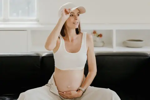 Kobieta w ciąży, w białym topie i czapce, siedzi na czarnej kanapie, czując uderzenia gorąca.
