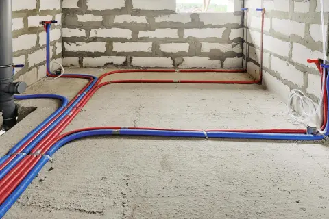 Nowa instalacja wodna w domu, z czerwonymi i niebieskimi rurami biegnącymi po betonowej podłodze i ścianach z bloczków.