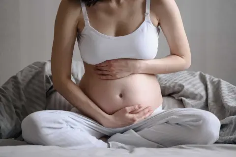 Gravid kvinna sitter på sängen med händerna på magen. Hon känner sig lite dålig i magen, en vanlig känsla under graviditeten.