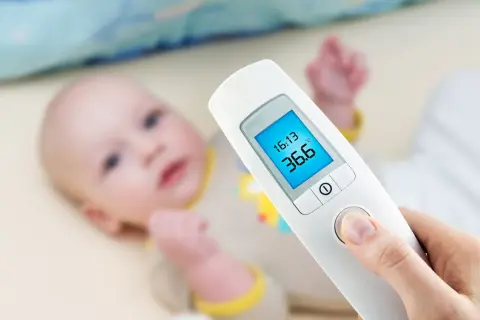 Termometr dla niemowlaka pokazuje 36.6°C. Dziecko leży w tle.