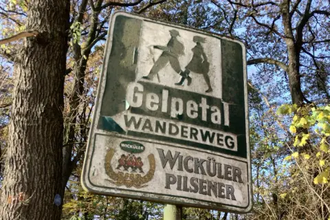 Schild am wanderparkplatz Gelpetal weist den Weg. Silhouetten von Wanderern und Bierwerbung.