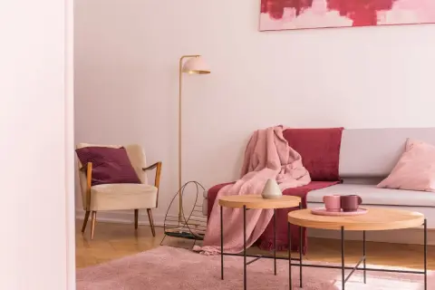 Przytulny salon z fotelem w kolorze ecru i amarantową poduszką. Na kanapie narzuta w tym samym amarantowym kolorze.