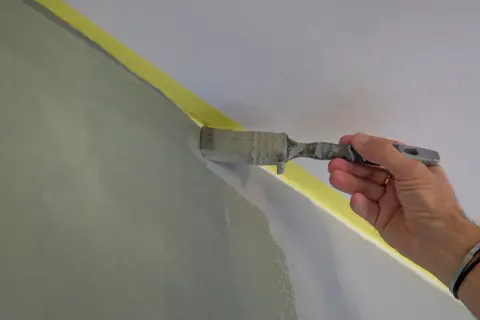 En hand målar en vägg inomhus med en pensel. Gul maskeringstejp skyddar taket.