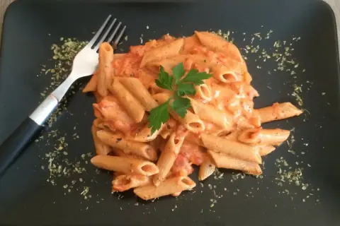 Penne w kremowym sosie pomidorowym z mascarpone, udekorowane natką pietruszki. Obok widelec i posypka z ziół.