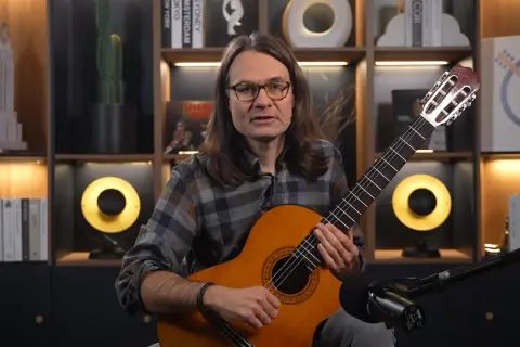 Mężczyzna z gitarą uczy, jak nauczyć się nut. W tle półki z książkami i dekoracjami.