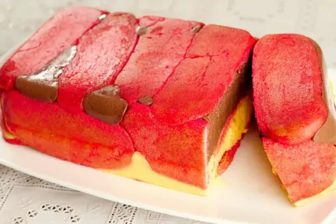 Una fetta di zuppa inglese tradizionale, con strati di pan di Spagna rosa, crema pasticcera gialla e cioccolato, decorata con savoiardi rossi e gocce di cioccolato.
