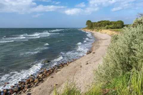 Windige Ostsee bei Berlin. Wellen brechen an einem steinigen Strand, dahinter eine sandige Steilküste mit Bäumen.