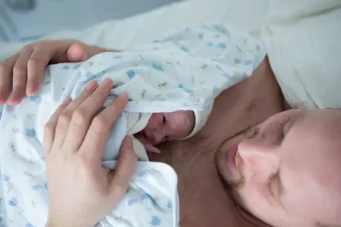 Pierwsze kangurowanie po cc. Ojciec tuli noworodka na swojej klatce piersiowej, otulając go w kocyk z motywem słoni.