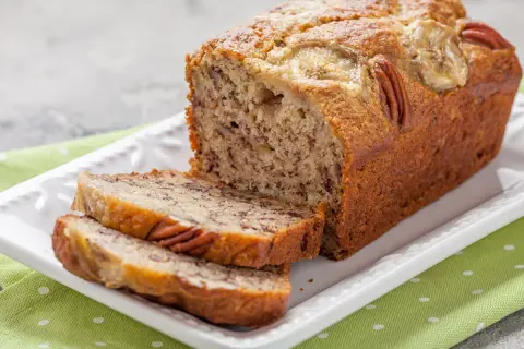 Pyszny chlebek bananowy z 2 bananów, z orzechami pekan i plasterkami banana na wierzchu, podany na białej paterze.