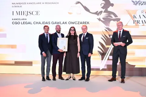 Pięć osób w strojach formalnych stoi na scenie, odbierając nagrodę.