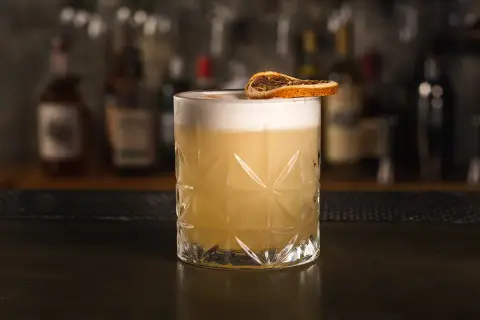 Klasyczny whiskey sour przepis: orzeźwiający koktajl z pianką i suszoną pomarańczą na tle barowych butelek.