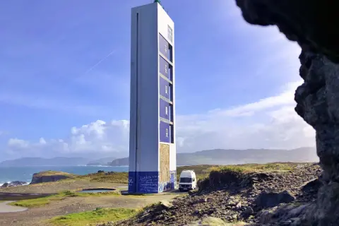 El faro de Punta Frouxeira se alza imponente sobre la costa, con un cielo azul y nubes blancas. Una furgoneta blanca descansa a su lado.