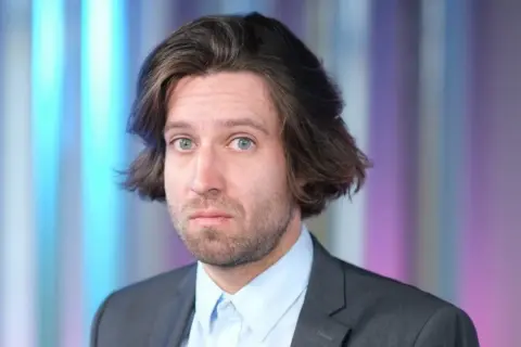 Michał Kempa stand up, w garniturze i jasnej koszuli, z burzą brązowych włosów i niebieskimi oczami, na tle kolorowych pasów.