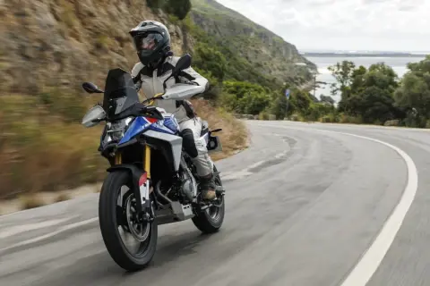 Motociclista en una maxi trail recorre una carretera sinuosa con vistas al mar y montañas verdes.