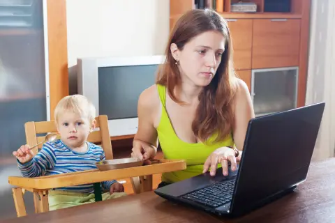 Matka z dzieckiem przy stole, zastanawia się, ile trwa urlop macierzyński, pracując na laptopie.