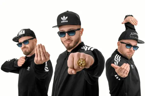 Trzech mężczyzn w czarnych bluzach z paskami Adidas i czapkach z daszkiem, pozujących jak zespoły disco polo.