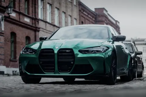 Zielony BMW M3 Competition z charakterystycznym grillem, zaparkowany na brukowanej ulicy przed ceglanymi budynkami.
