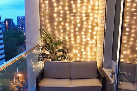 Przytulny balkon w bloku z sofą i palmą, ozdobiony ciepłym oświetleniem.