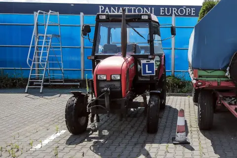 Czerwony traktor z literą "L" (kategoria t prawo jazdy) stoi obok przyczepy. W tle niebieska ściana i drabina.