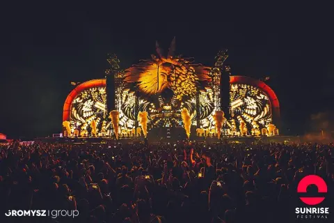 Nocny hardstyle festival z ogromną sceną i tłumem ludzi. W tle świecąca, złota głowa w koronie.