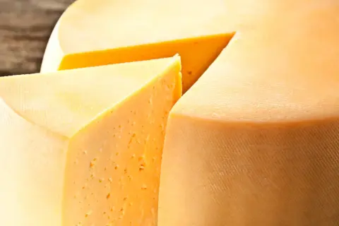 Kawałek sera Gouda z widocznymi oczkami i fragmentem całego kręgu sera.