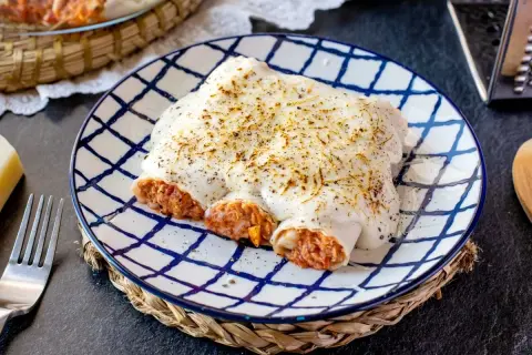 Canelones de atún y huevo gratinados, cubiertos de bechamel y queso, servidos en plato a cuadros.