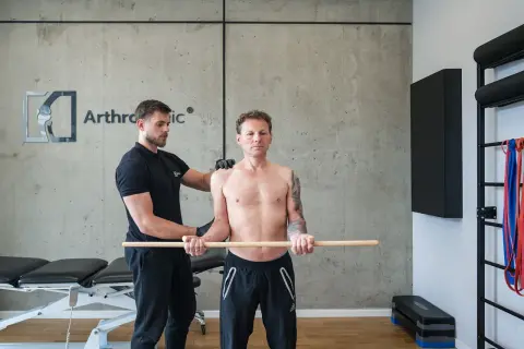 Fizjoterapeuta pomaga pacjentowi w ćwiczeniach rehabilitacja barku po operacji. Mężczyzna trzyma kij, wykonując ruchy ramionami.