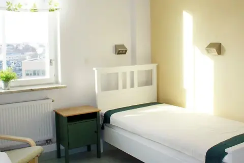 Ett ljust rum med en vit säng, grön sänggavel och en grön nattduksbord. Fönstret visar en stadsvy. Känns som ett rum där någon är inlagd på psyk.