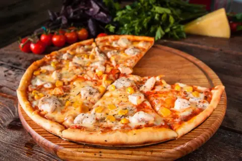 Pyszna pizza z kurczakiem i kukurydzą, podana na drewnianej desce. Obok świeże pomidorki i zioła.
