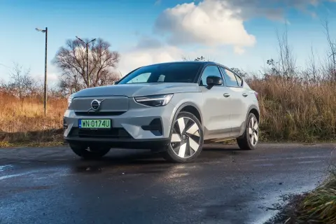 Szary Volvo C40 na mokrej drodze, otoczony suchą trawą i drzewami.