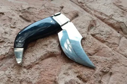 Navaja capaora de mango negro y hoja curva, lista para la acción sobre una superficie rocosa.