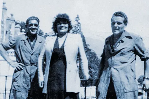 Trois personnes posent devant un paysage. L'homme de gauche, peut-être le fondateur Millet, sourit.