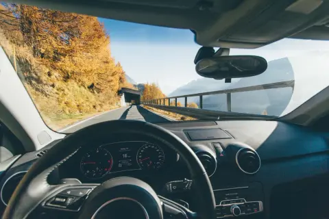 Volant d'une voiture en mouvement, tableau de bord affichant 16 km/h. Paysage d'automne en Italie, arbres dorés, montagnes et tunnel.