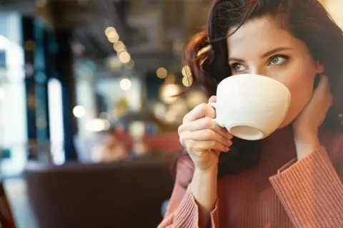 En kvinna med mörkt hår dricker kaffe ur en vit kopp. Hon har en stickad tröja och sitter på ett café. Kanske lite kaffe på tom mage för att vakna till.