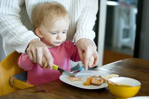 Mama pomaga dziecku kroić rybę na talerzu. To idealne pancakes dla niemowlaka, które uczy się jeść samodzielnie.