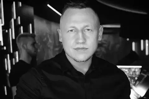 DJ Iroon w czerni, gotowy do rozkręcenia imprezy. W tle neonowe światła klubu.