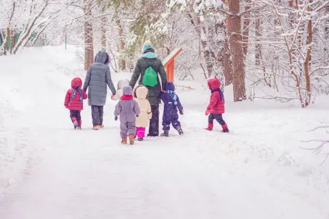 Schneebedeckter Weg im Wald. Eine Gruppe Kinder und eine Erzieherin gehen zum Kindergarten.