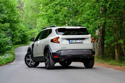 Nowy Dacia Duster 3 na krętej leśnej drodze. Nowoczesne światła i masywna sylwetka podkreślają jego terenowy charakter.