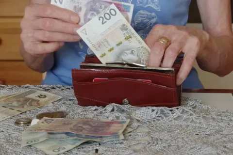 Dłonie wkładają banknot 200 zł do portfela. Na stole leżą inne banknoty i monety, co skłania do refleksji, ile wynosi średnia emerytura.