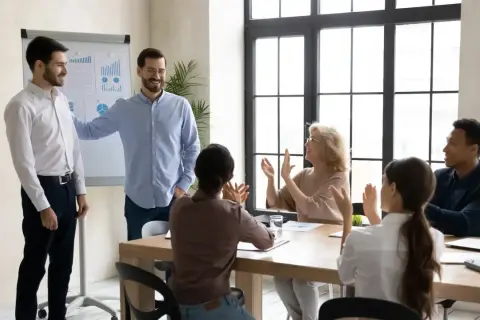 Dobry kierownik docenia zespół. Mężczyzna gratuluje pracownikowi, który prezentuje wykresy, a reszta zespołu klaszcze.