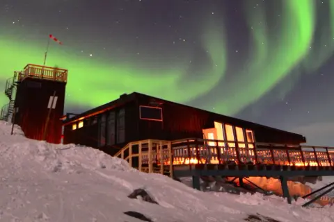 En snöig sluttning leder upp till en mysig aurora sky station. Norrskenet dansar grönt över stjärnorna.