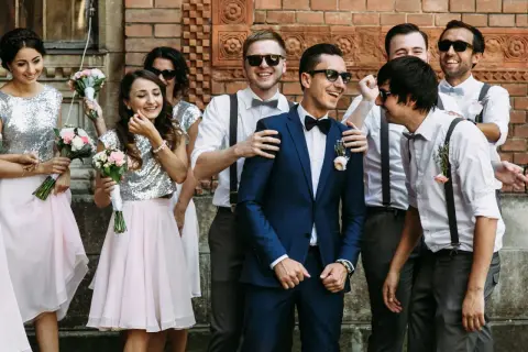 Uśmiechnięci druhny i drużbowie pozują do zdjęcia. Dziewczyny w błyszczących topach i różowych spódniczkach trzymają bukieciki. Panowie w garniturach i z szelkami wyglądają elegancko.