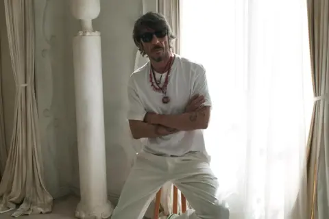 Pierpaolo Piccioli, con occhiali da sole e collane, posa con le braccia conserte in un ambiente chiaro con tende e colonne.