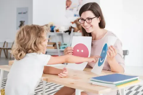 Nauczycielka z uśmiechem pokazuje dziecku karty z buźkami, wspierając jego przygotowanie pedagogiczne.