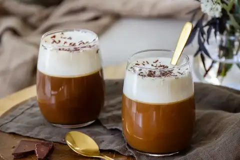 Två glas med kaffe drinkar med alkohol, toppade med grädde och chokladströssel. En gyllene sked vilar i det ena glaset.