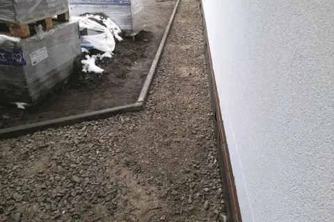 Ścieżka z tłucznia drogowego obok białej ściany budynku i palet z materiałami budowlanymi.