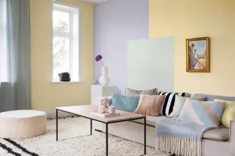 Ett vardagsrum med pastell inredning. Ljusgula, ljuslila och ljusgröna väggar skapar en mjuk atmosfär. En beigefärgad soffa är prydd med kuddar i olika pastellfärger och mönster.