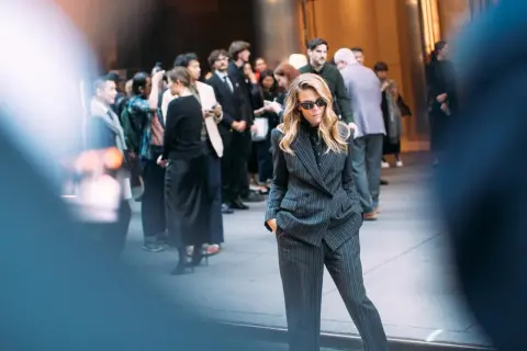 Eine Frau in einem gestreiften Anzug mit einer Hose, die an eine blaue Anzughose kombinieren Herren erinnert. Sie steht im Vordergrund, umgeben von einer Menge.