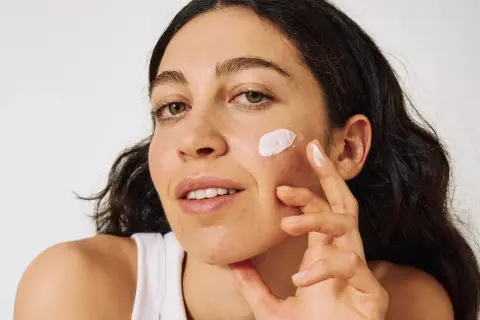 Frau trägt sanft Sonnencreme auf ihre Wange auf, um ihre Haut vor Akne zu schützen.