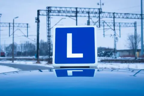 Niebieska tablica z literą "L" na dachu samochodu, symbolizująca naukę jazdy i przyszłe testy na kierowcę.